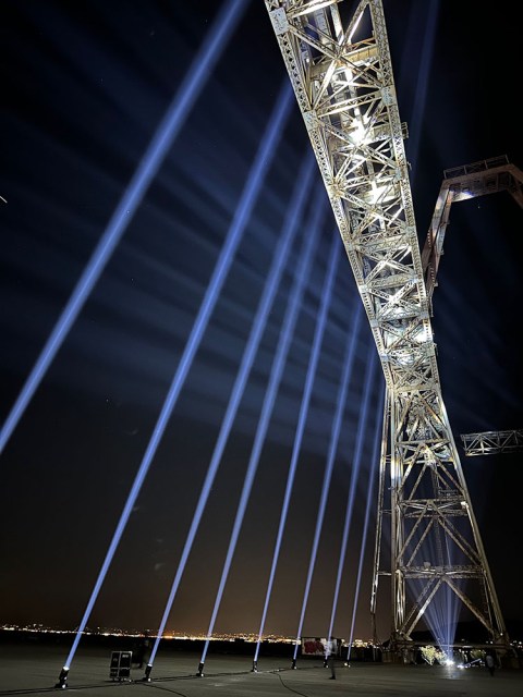 Tall metal framework lit by spotlights at night, with multiple beams of blue light projecting upward into the sky alongside the structure. City lights are visible in the background.