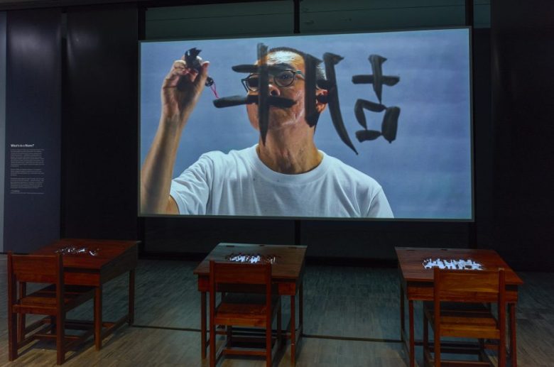 A video projection shows a person writing large calligraphy on glass; three wooden desks with small objects are arranged in front of the screen.