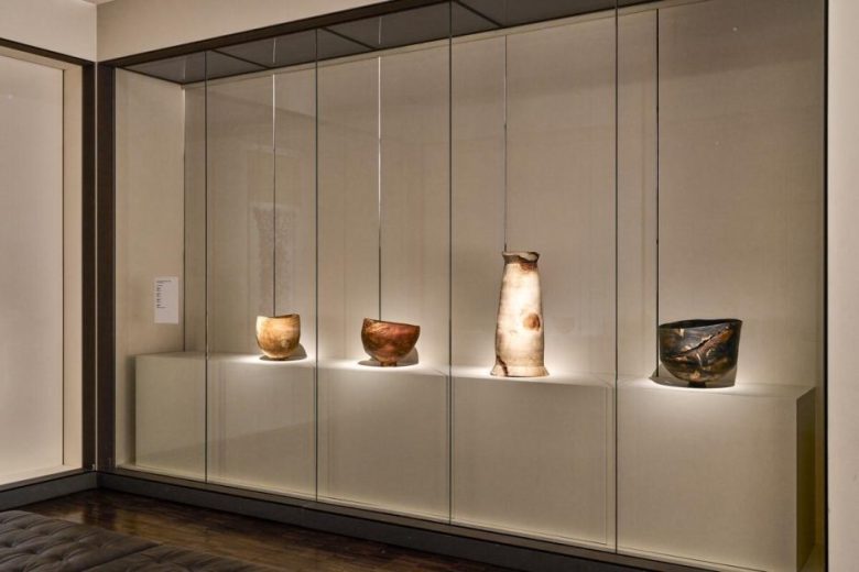 Four ceramic vessels of varying shapes and colors are displayed on a white shelf inside a glass case in a modern, well-lit museum or gallery setting.