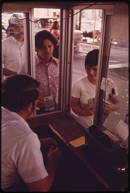Three people stand at a service window, presenting identification to an attendant seated inside a booth. Signs and paperwork are visible on the window and desk, creating a distinctly anthropic scene of human interaction and administrative routine.