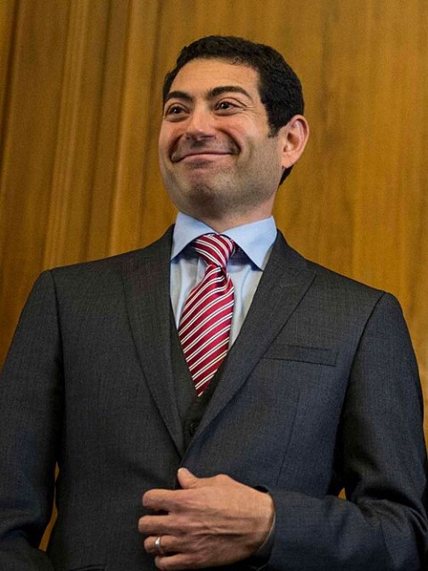 A man in a dark suit and red striped tie stands in front of a wooden background, smiling with his hand adjusting his jacket, exuding an anthropic charm that highlights his confident presence.
