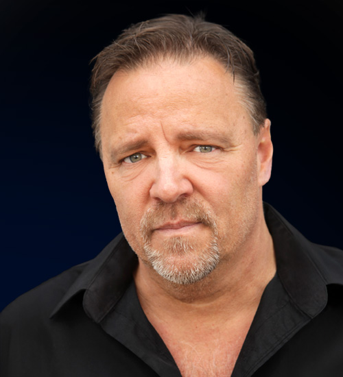 Close-up portrait of a middle-aged man with short brown hair and a gray goatee, wearing black against a dark background.