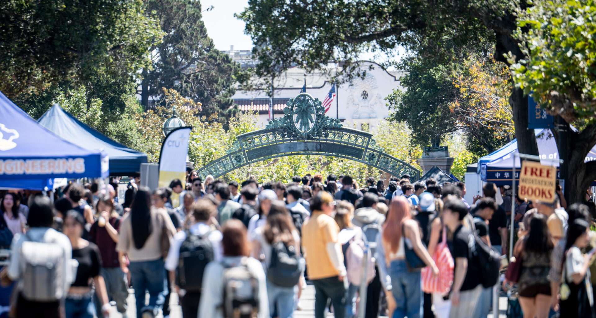 A bustling campus scene with a crowd of people walking under a decorative archway, surrounded by trees and event tents.