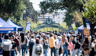 A bustling campus scene with a crowd of people walking under a decorative archway, surrounded by trees and event tents.