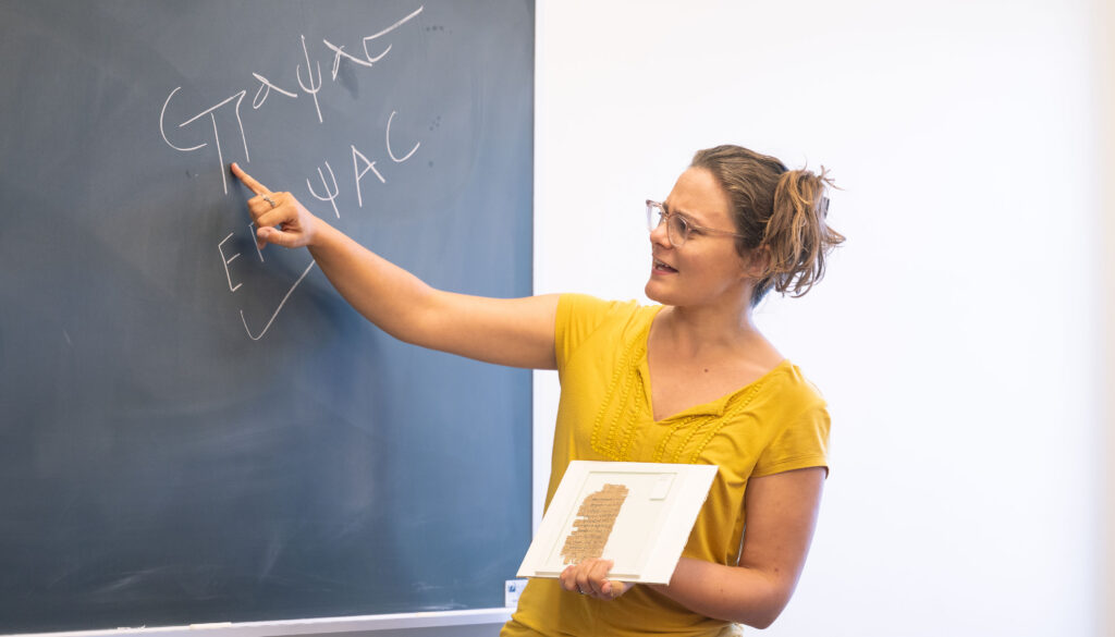 a woman in mustard-colored blouse pointing at Greek letters on a chalkboard