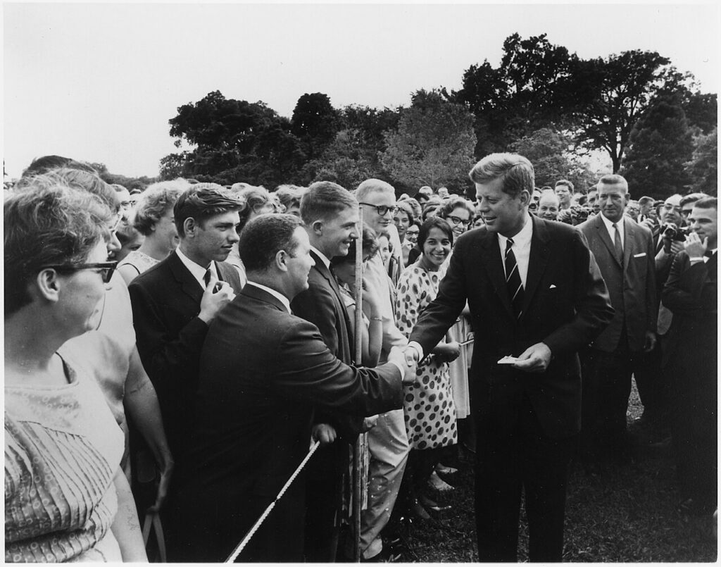 A black and white image showing President John F. Kennedy surrounding by people, shaking a Peace Corps volunteer's hand.
