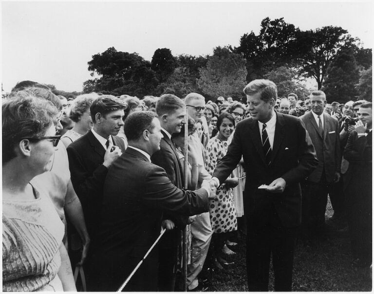 A black and white image showing President John F. Kennedy surrounding by people, shaking a Peace Corps volunteer's hand.