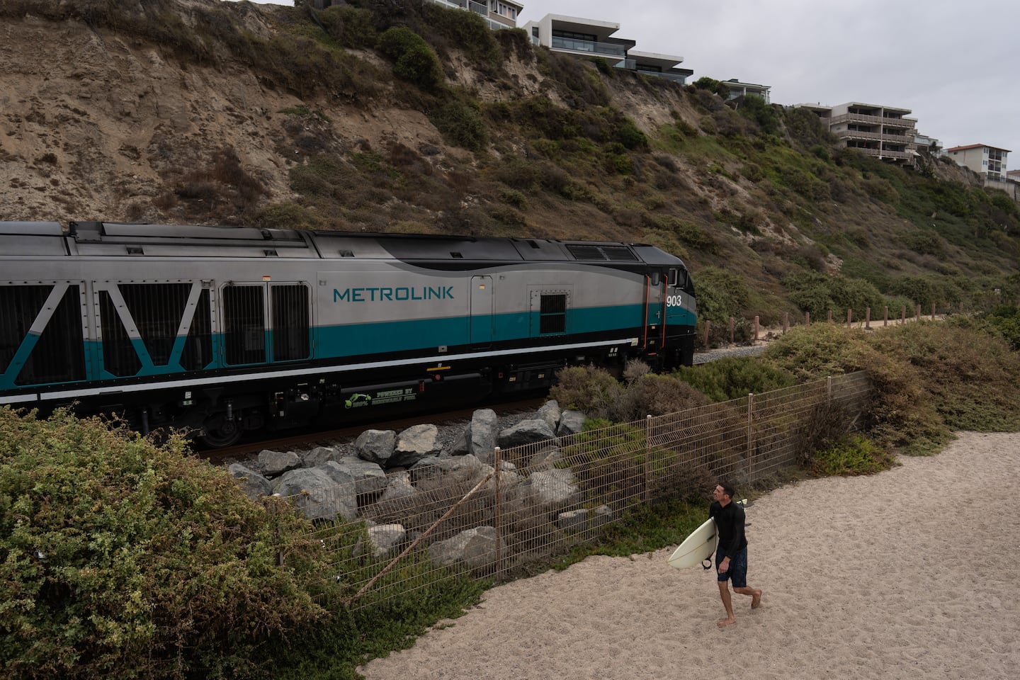 A surfer walked along North Beach as a Metrolink train traveled along the coast in San Clemente on Sept. 18.