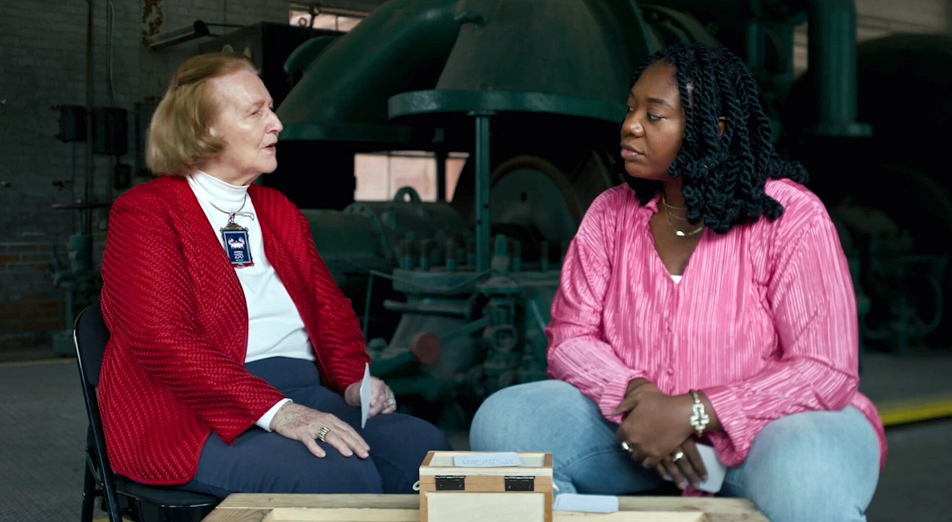 An older white woman at right, near 80 years old, speaks with a thoughtful expression while a younger Black woman, near 30, listens.