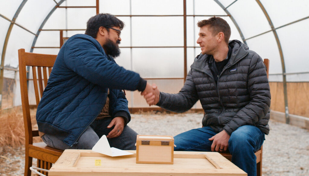 Two men wearing light winter jackets are sitting under a plastic canopy with dry winter fields visible behind them. The man of color on the left and the white man on the right are shaking hands.