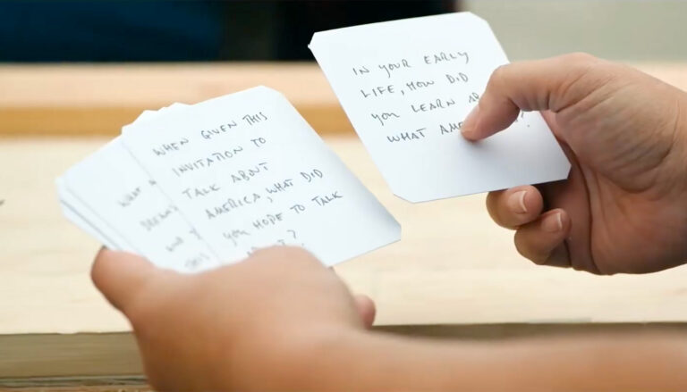 Two hands hold several white cards with handwritten questions on them. One of the cards says, "When given this invitation to talk about America, what did you hope to talk about?"