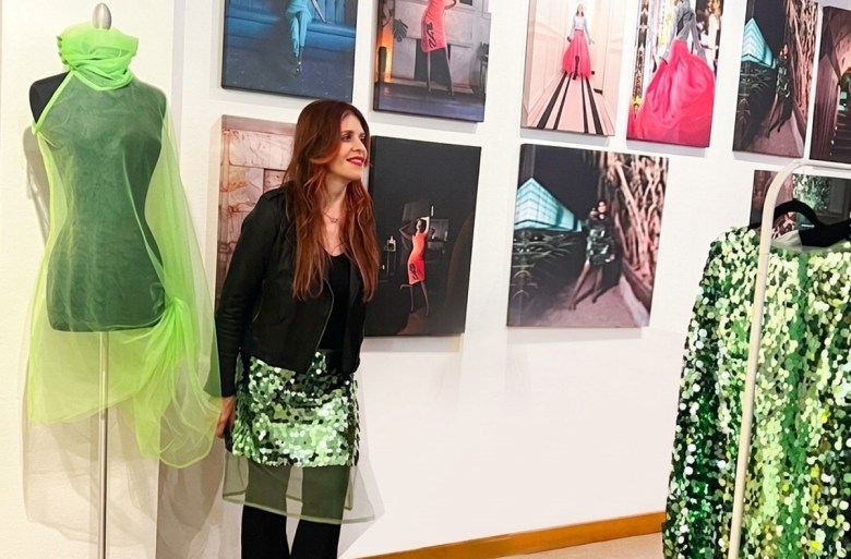 A fashion designer stands next to a green-draped mannequin in a fashion exhibit, wearing a black leather jacket and a green sequin skirt, with fashion photos displayed on the wall behind.