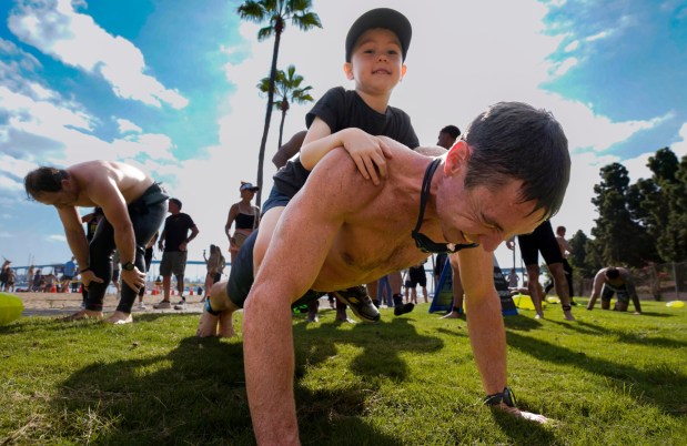 CORONADO, CA - SEPTEMBER 19, 2026: Nate Middleton of Eastlake in Chula Vista, does push-up while his four-year-old son, Finn Middleton, sits on his back back, during the 6th annual Swim for SOF ( Special Operations Forces), put on by the Honor Foundation, at Tidelands Park, during a stop on the way during the 3.2 mile swim from Glorietta Bay Park to the USS Midway Museum, Saturday September 19, 2026, in Coronado, California. (Howard Lipin / For The San Diego Union-Tribune)