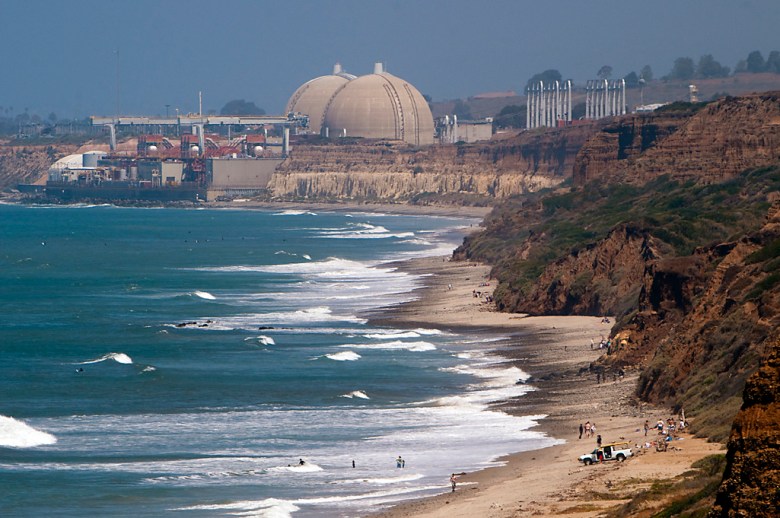 Turquoise waves roll onto a sandy California coast below rugged cliffs, with beachgoers and a large domed power plant in the background.