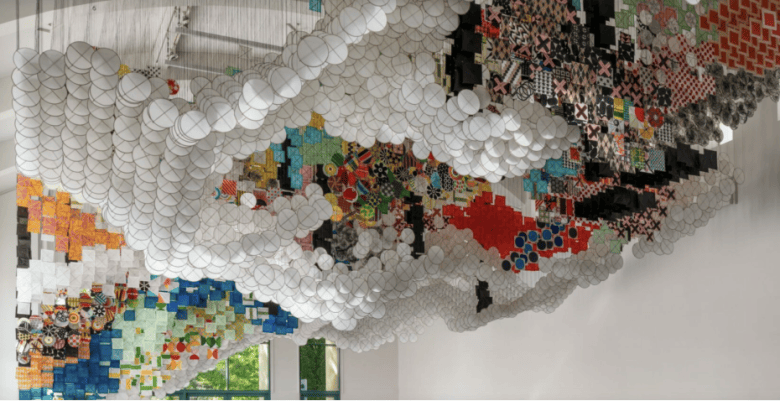 Colorful, geometric collage art installation covers the ceiling, featuring paper discs, fabric squares, and various patterns suspended above gallery space. Large windows let in natural light.