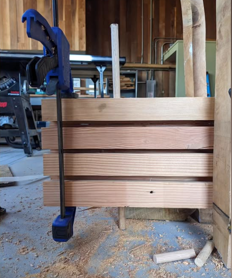 A close-up view of four thick, rectangular wooden blocks stacked horizontally with small gaps between them in a woodworking workshop. A large blue and black bar clamp is attached vertically on the left side, holding the stacked wood securely together. A vertical wooden dowel projects upward from the center top of the stack. In the background, sawdust and small dowel offcuts are on the floor, while workshop equipment, including a table saw, stands in the softly blurred background.
