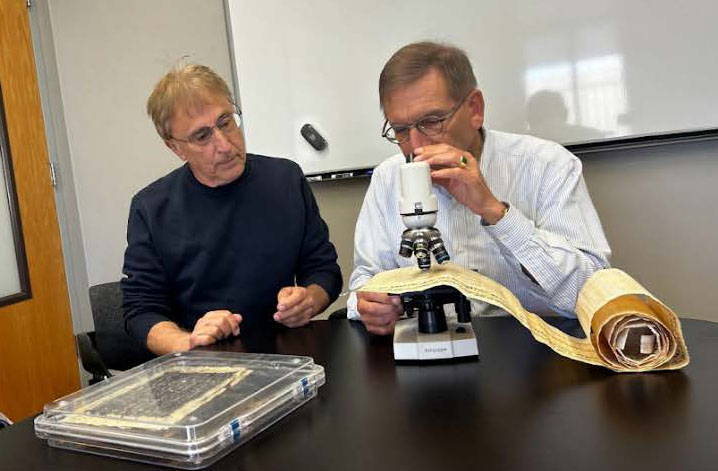 two men examining an unrolled papyrus under a microscope