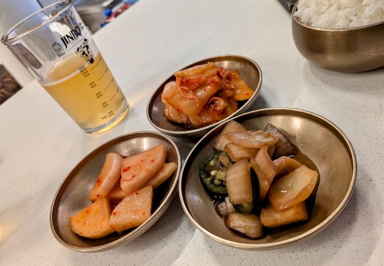Three small dishes of Korean side dishes, a bowl of white rice, and a glass of beer on a white tabletop.