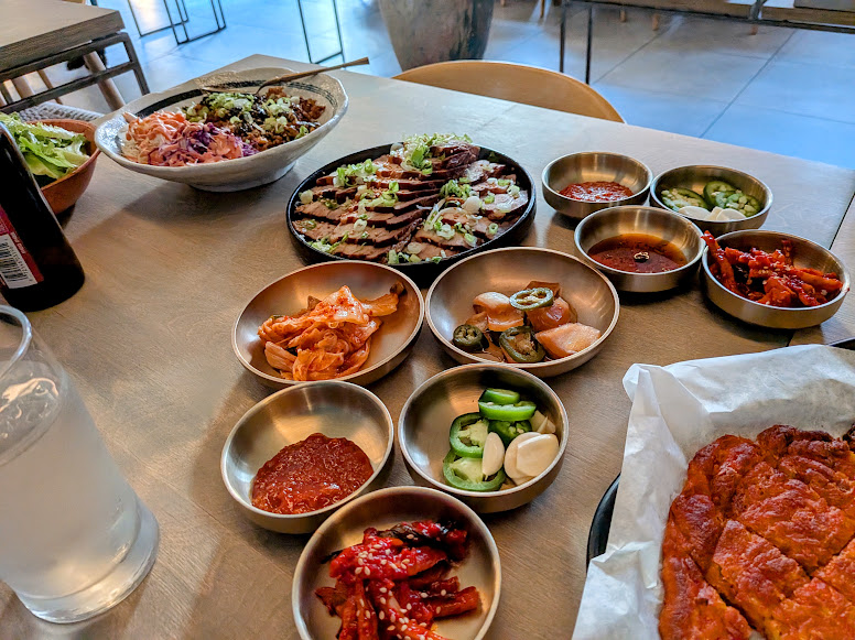 A table set with Korean dishes, including sliced meat, assorted banchan in small bowls, a large salad, a glass of water, and a partially visible fried dish.