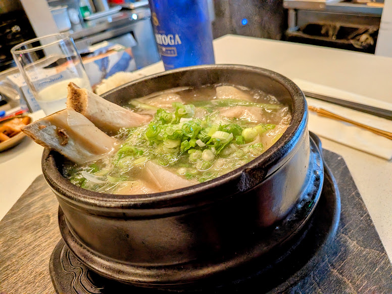 A hot stone bowl of soup with beef bones, green onions, and glass noodles sits on a table, with chopsticks and a drink in the background.