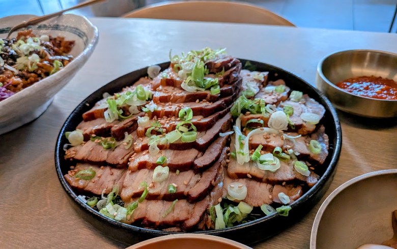 A large plate of sliced cooked beef topped with chopped green onions, surrounded by side dishes and a small bowl of red sauce.