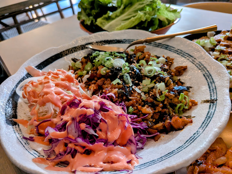 A plate with seasoned chopped meat, sliced green onions, shredded red cabbage, and white cabbage drizzled with pink sauce, with a side of lettuce in the background.