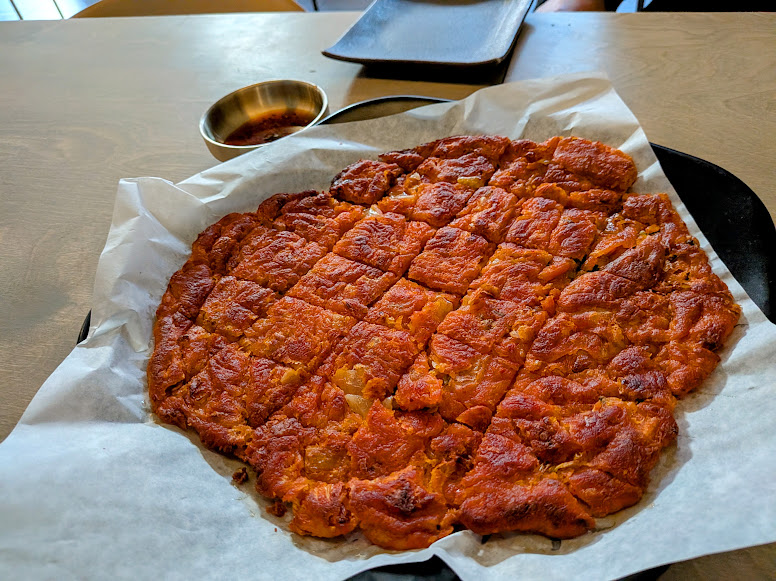 A round, sliced kimchi pancake sits on parchment paper, with a dipping sauce in a small metal bowl nearby on a wooden table.