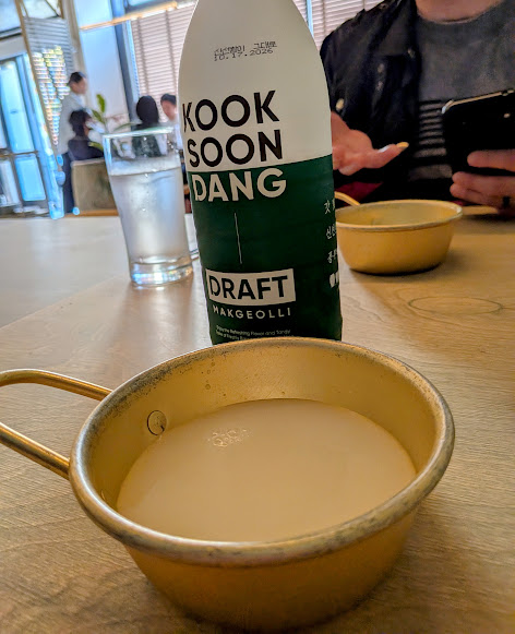 A gold bowl filled with makgeolli sits on a table beside a bottle labeled “Kook Soon Dang Draft Makgeolli.” A glass of water and a person holding a phone are in the background.