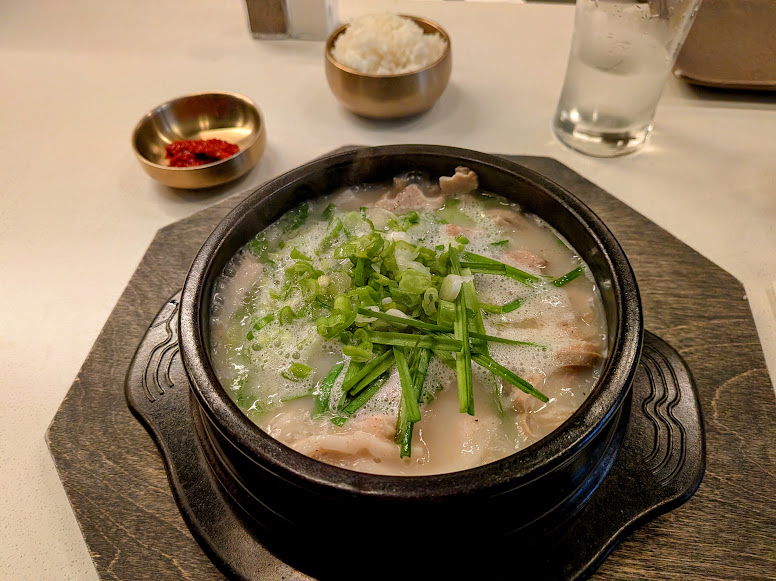 A bowl of Korean soup with sliced meat and green onions, served with a side of rice, red chili paste, and a glass of water on a wooden tray.