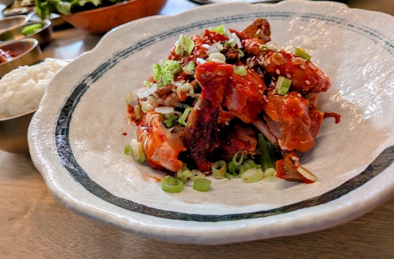 A plate of spicy marinated octopus topped with chopped green onions and sesame seeds, served on a white dish with a side of rice visible in the background.