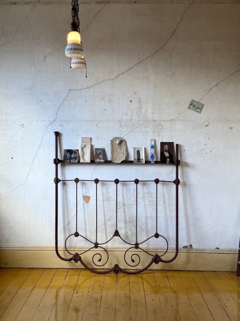 An old metal bed frame leans against a cracked wall with vintage photos and papers displayed on a shelf, beneath a single hanging light. The floor is wooden and glossy.