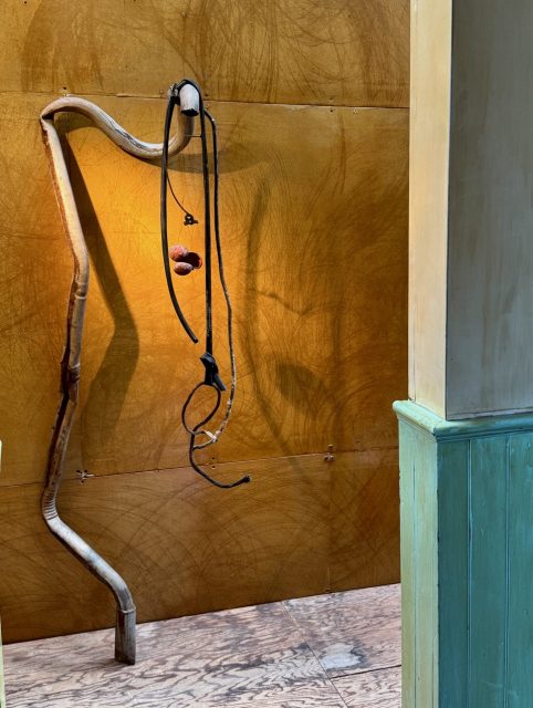 Curved metal pipes and black cables are mounted on a brown textured wall, with two reddish spheres suspended in the middle, next to a green paneled corner.