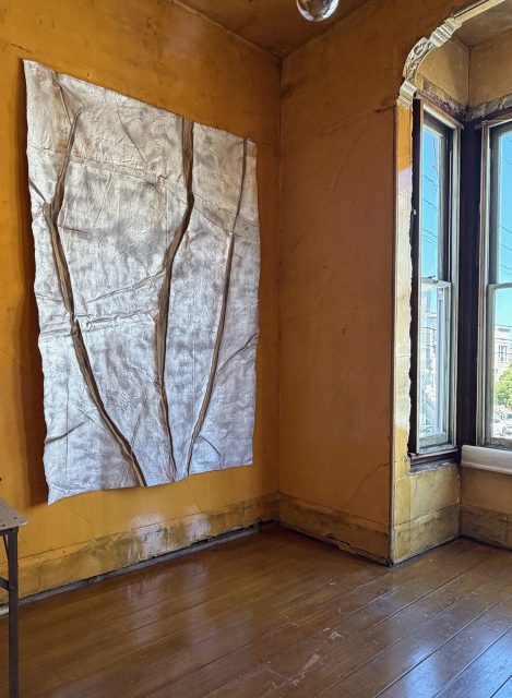 A textured metallic artwork hangs on a worn orange wall in a sunlit room with wooden floors and an open window.