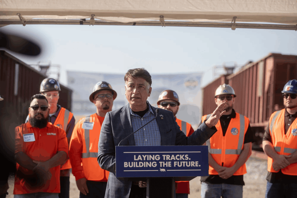 Man speaking at a podium between freight rail cars, with rail workers in orange safety gear behind him. The podium reads, “LAYING TRACKS. BUILDING THE FUTURE.”