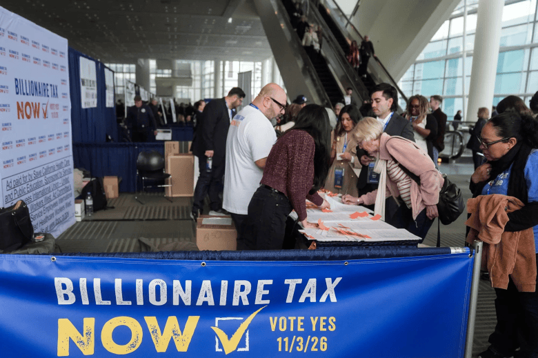 People sign papers at an information table beneath a blue “BILLIONAIRE TAX NOW” campaign banner inside a convention center.