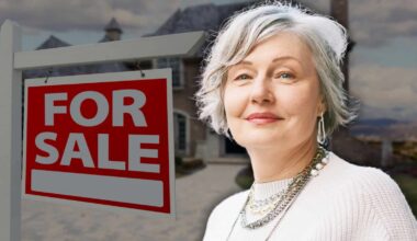 A smiling senior woman with gray hair, wearing a white sweater and layered necklaces, stands next to a red and white 'FOR SALE' sign. A large house with a dark roof is visible, blurred, in the background under a cloudy sky.