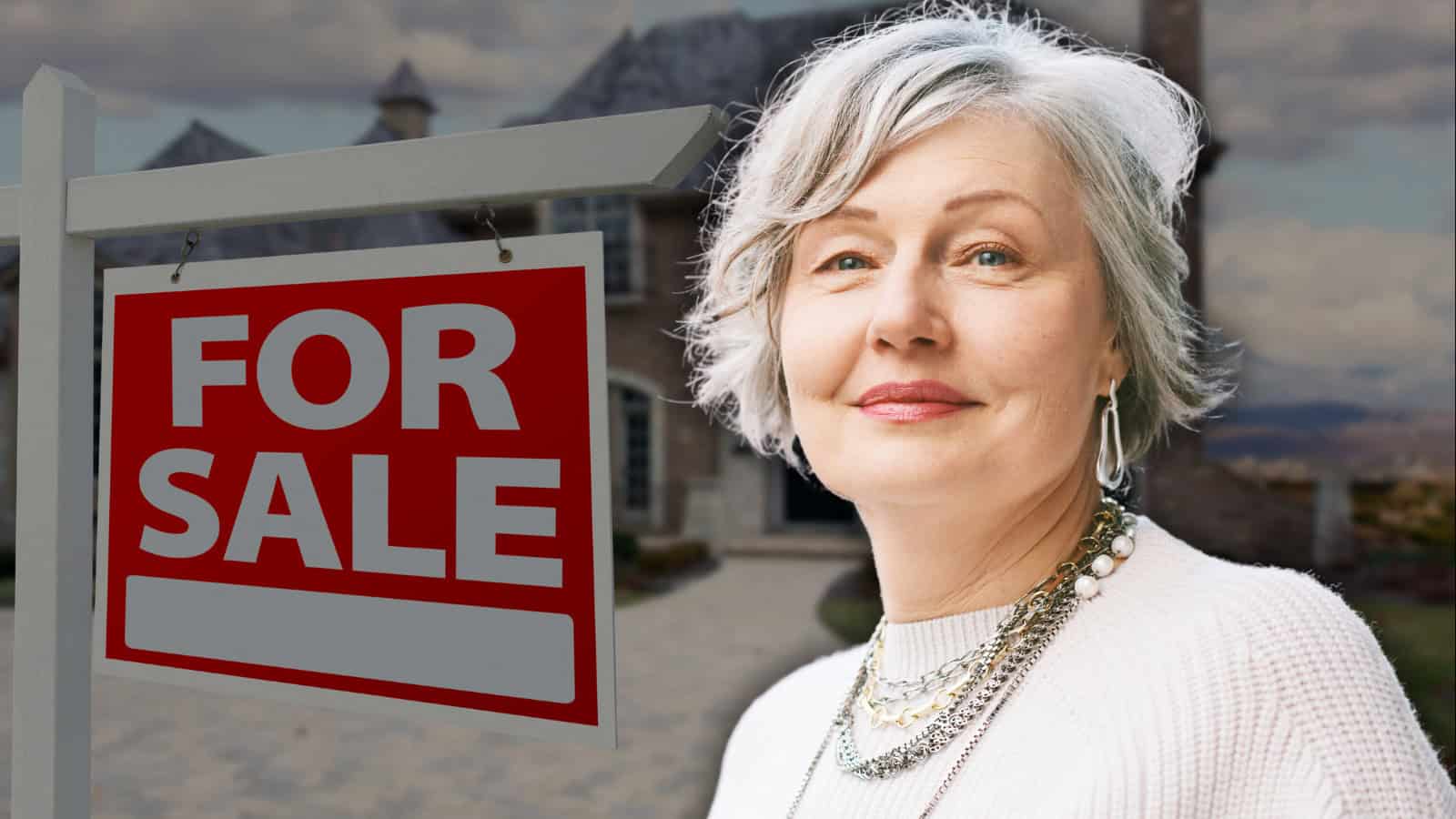 A smiling senior woman with gray hair, wearing a white sweater and layered necklaces, stands next to a red and white 'FOR SALE' sign. A large house with a dark roof is visible, blurred, in the background under a cloudy sky.
