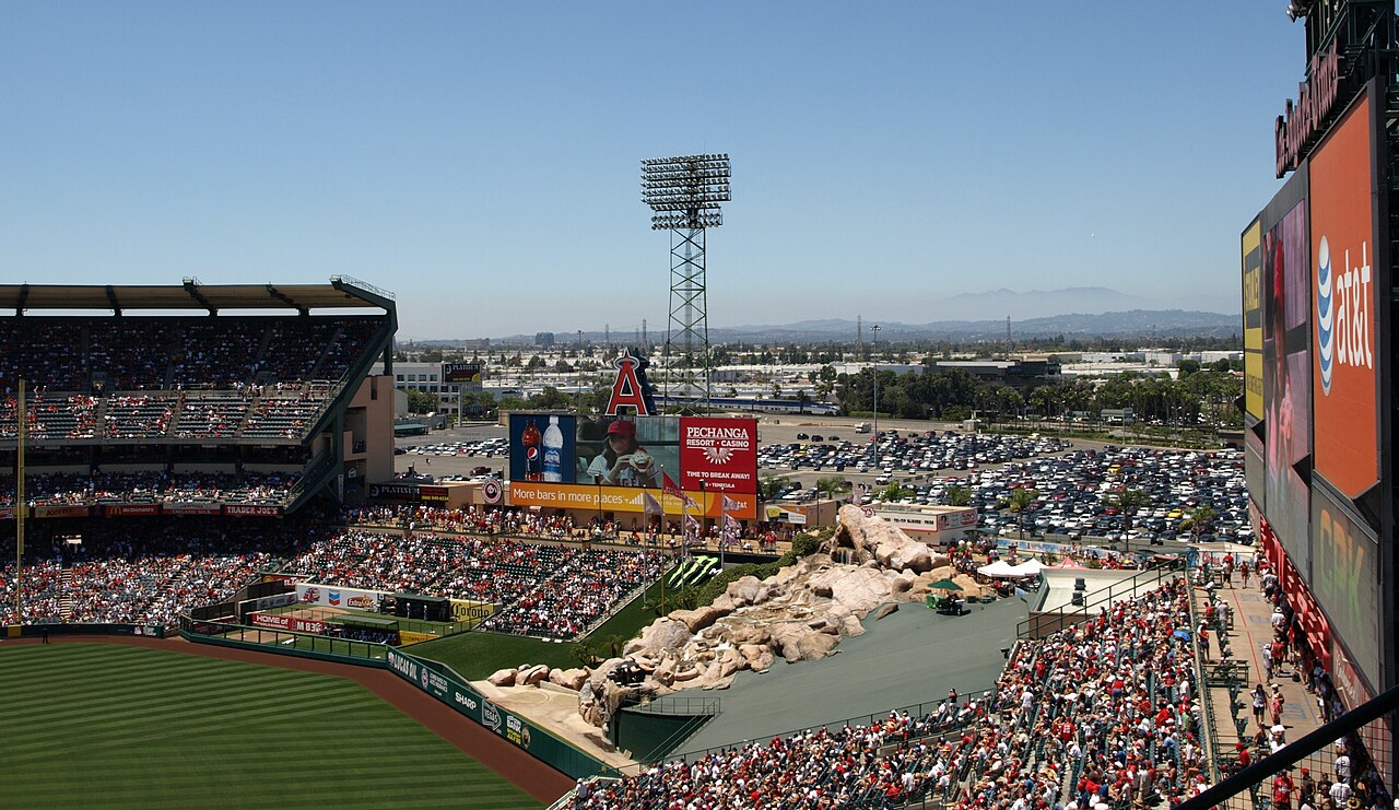 Angels baseball stadium with a packed crowd, large scoreboard, outfield rock feature, and distant view of mountains under a clear blue sky.