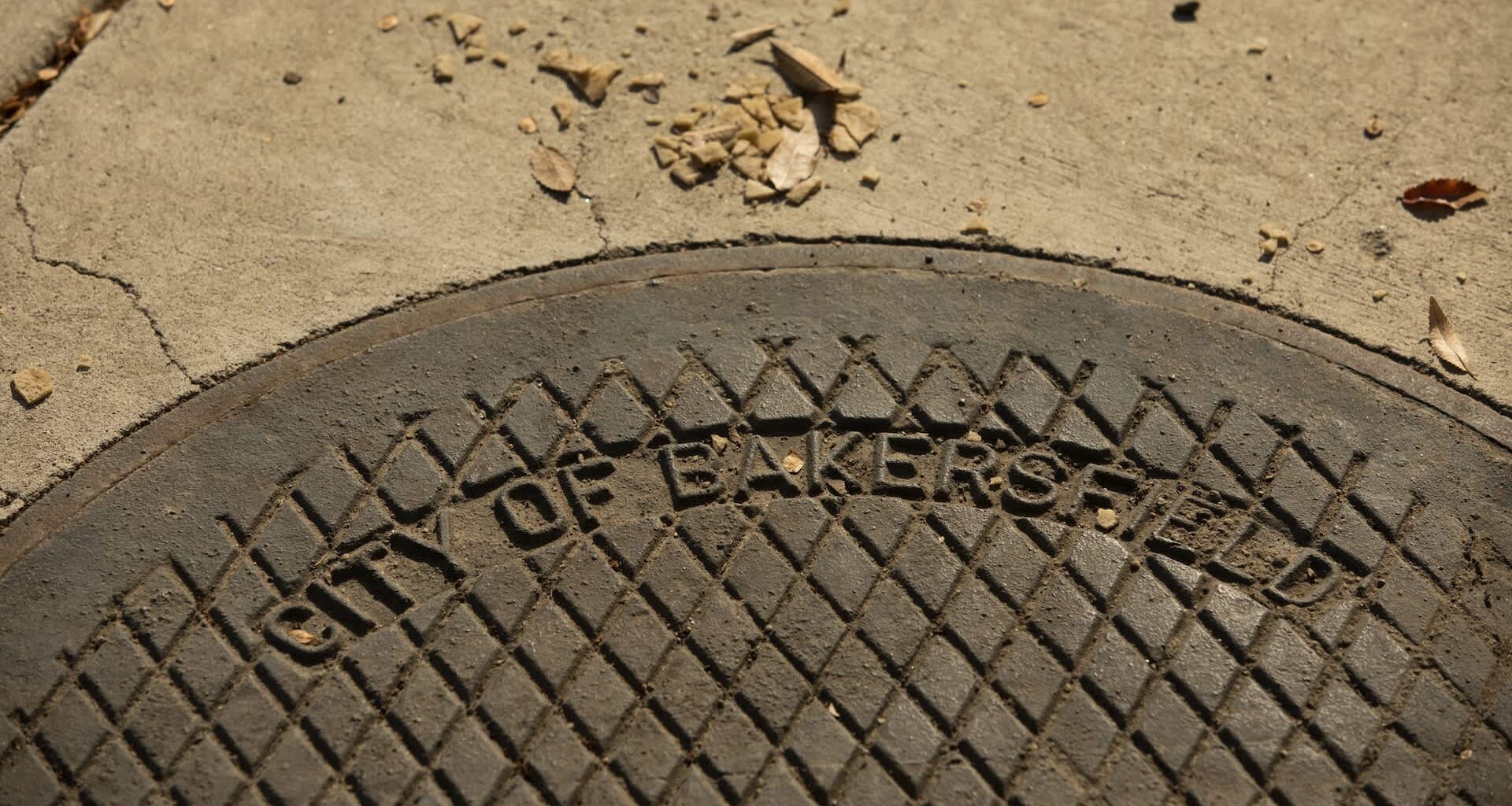 A manhole cover inscribed with "City of Bakersfield" on a concrete surface with scattered leaves and debris.