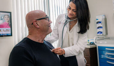 A physician in a white coat leans over a seated, older man while examining him.