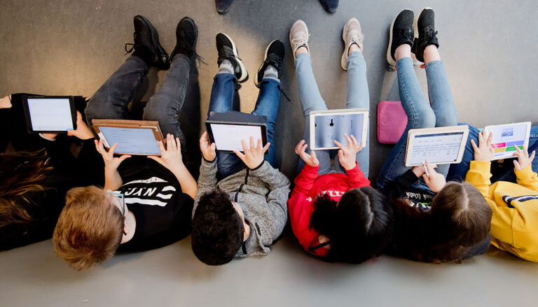 A photo looking down on six children, each of whom is engaging with the screen of a digital tablet.