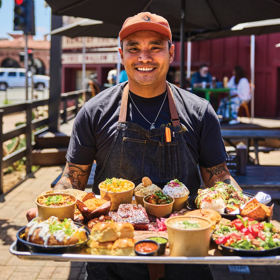 smiling chef holding a tray with a variety of colorful dishes outdoors, 25 restaurants defining california dining, heritage barbecue, san juan capistrano california, executive chef nicholas echaore serves a medley of side dishes