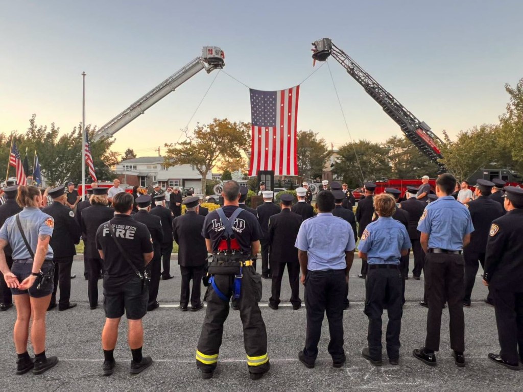 Firefighters at a 9/11 remembrance ceremony, with a large American flag suspended between two fire truck ladders.