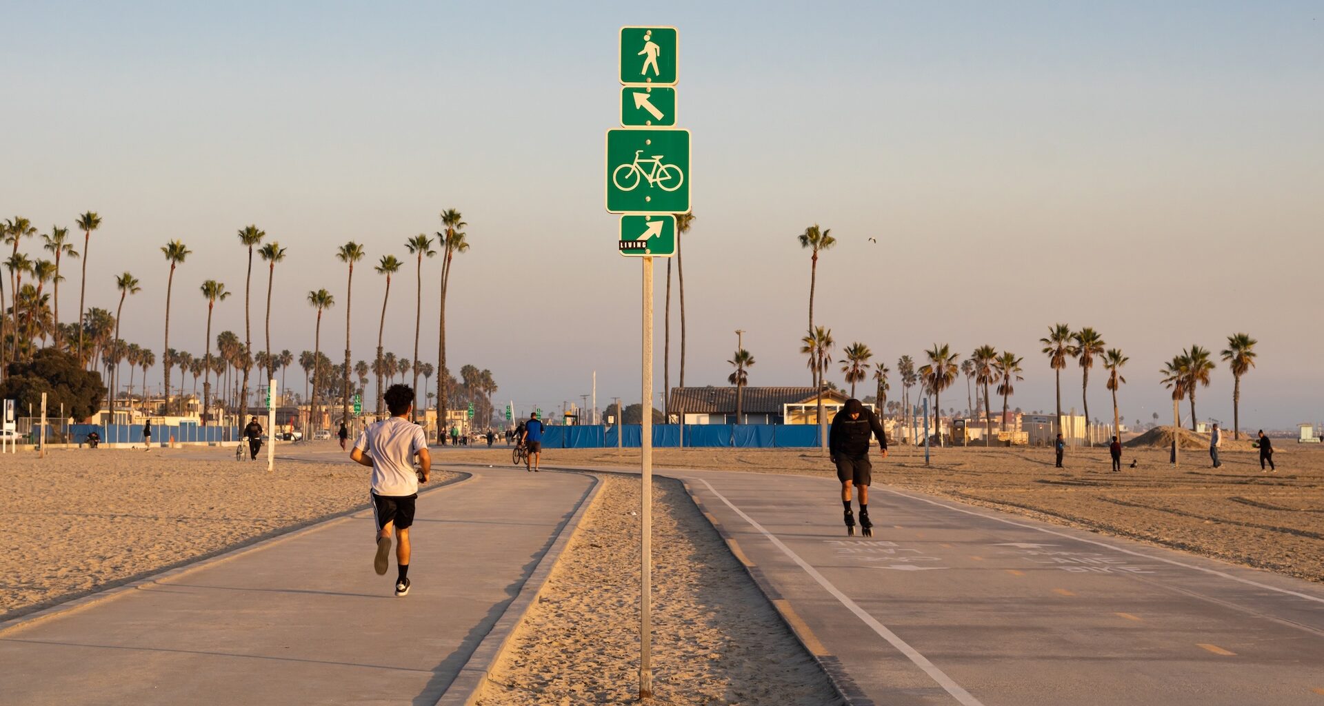 A pedestrian and a person on rollerblades along a beachside walkway with palm trees and signs for walking and biking.