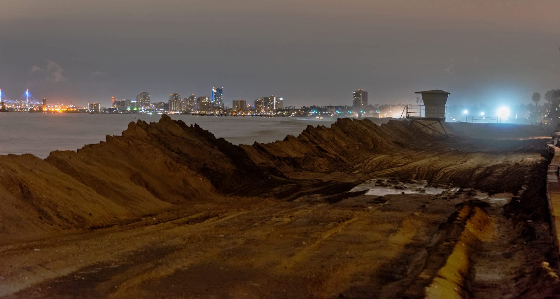 A sandy beach at night with city lights and a lifeguard tower in the distance.
