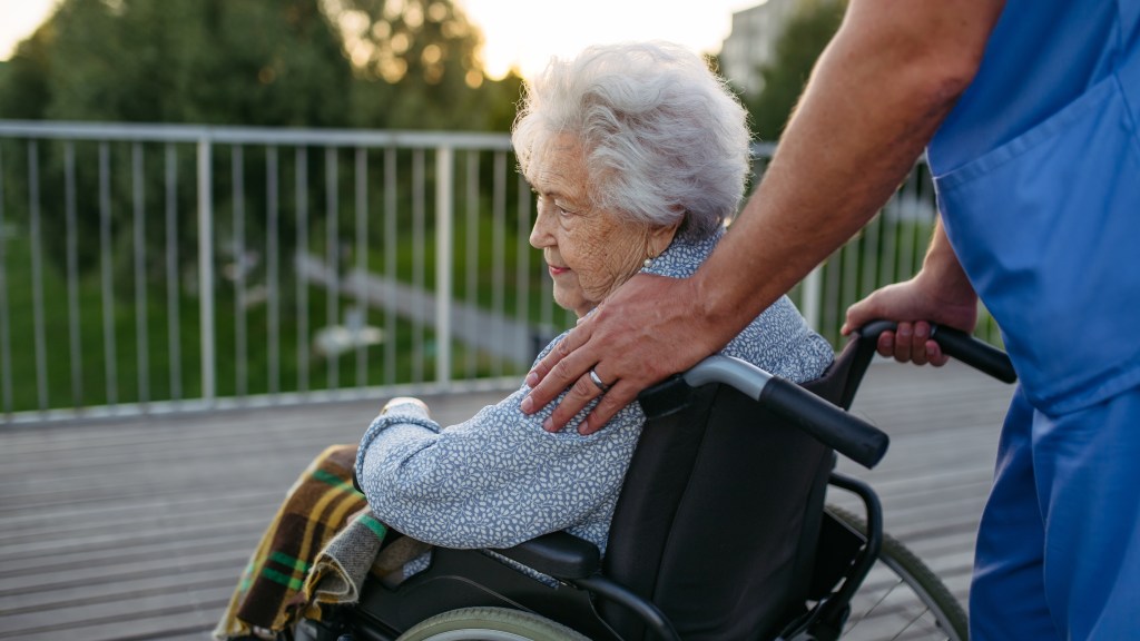 Male caregiver accompanying an elderly patient in a wheelchair on an outdoor walk.