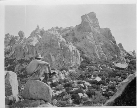 Black-and-white landscape shows a seated hiker in a wide-brimmed hat gazing at rugged granite peaks, boulders, and shrubs; serene, adventurous mood.