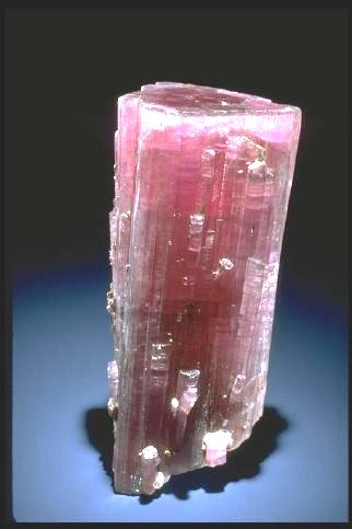 A tall, translucent pink mineral crystal stands upright on a dark surface, with vertical striations, rough edges, and sparkling reflective facets.