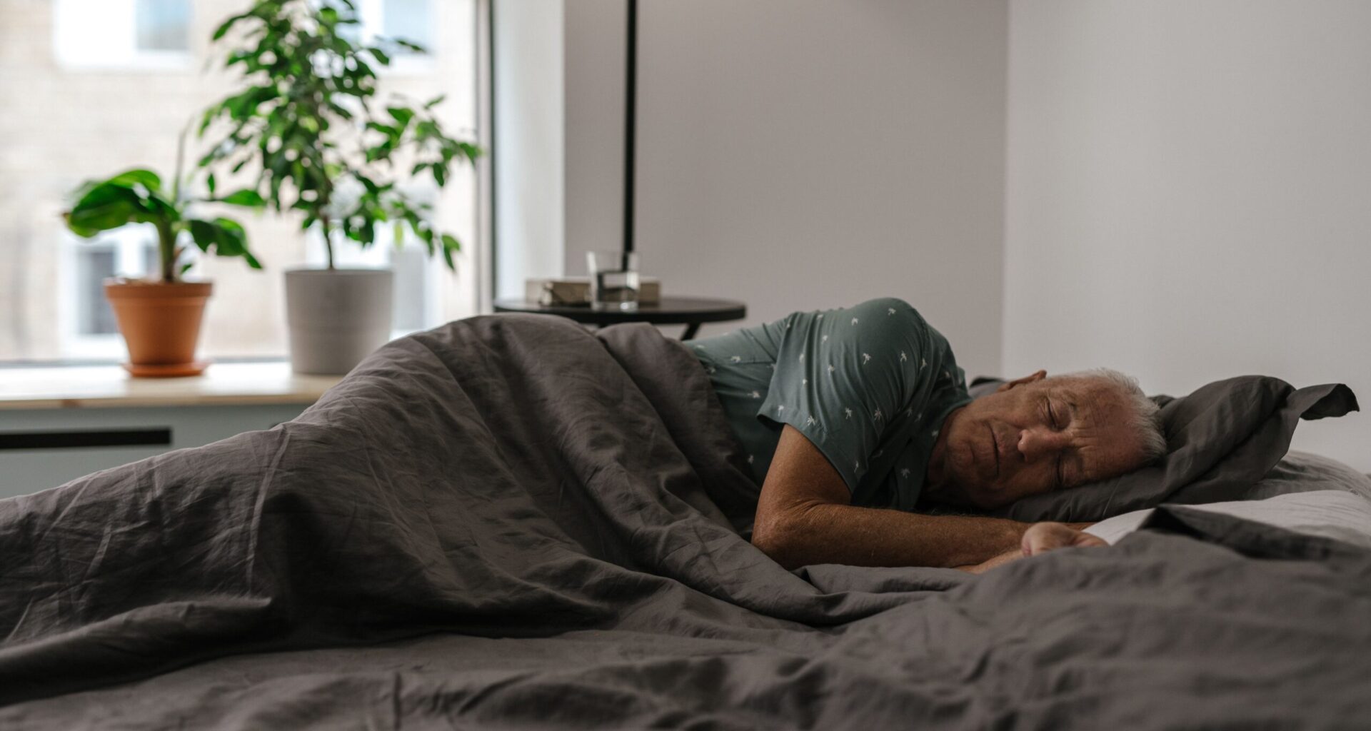 Senior man resting peacefully on a bed inside a room with natural light and plants.