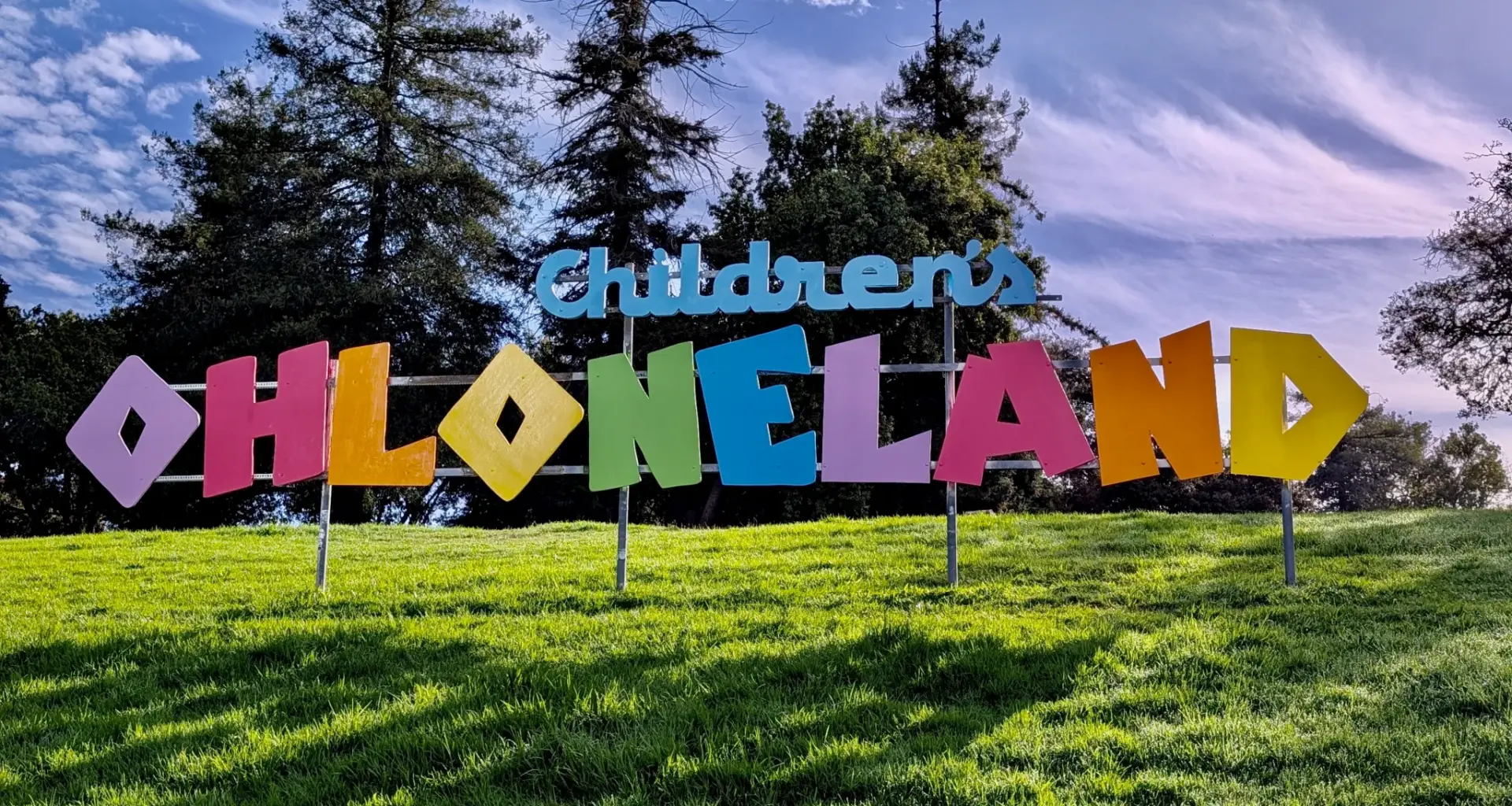 A rainbow-colored sign on a patch of grass under a puffy clouded sky and in front of tall pine trees reads Children's Ohloneland.